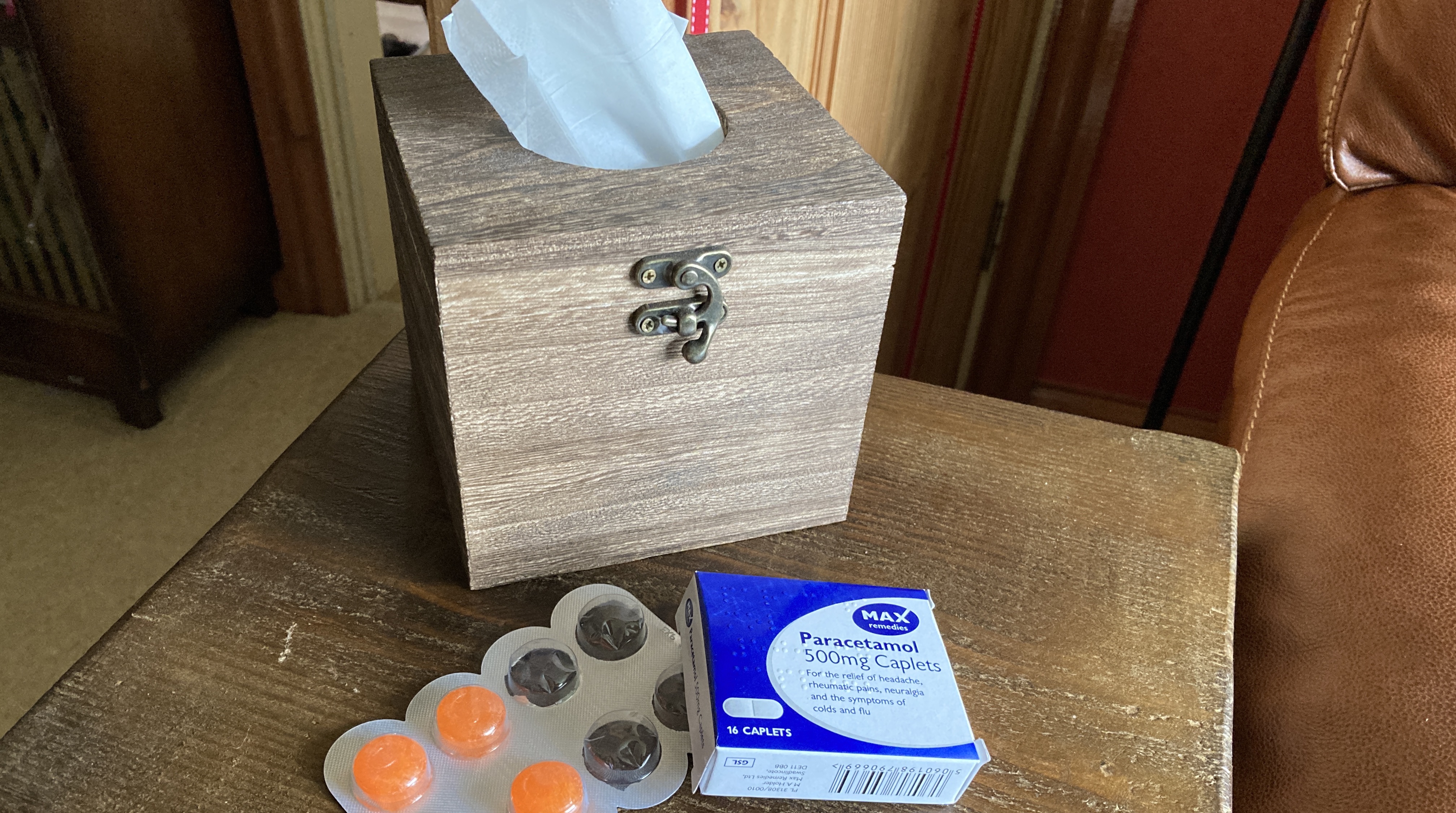 A wooden box of tissues, a packet of paracetamol and a strip of Strepsils lie on a small wooden table.