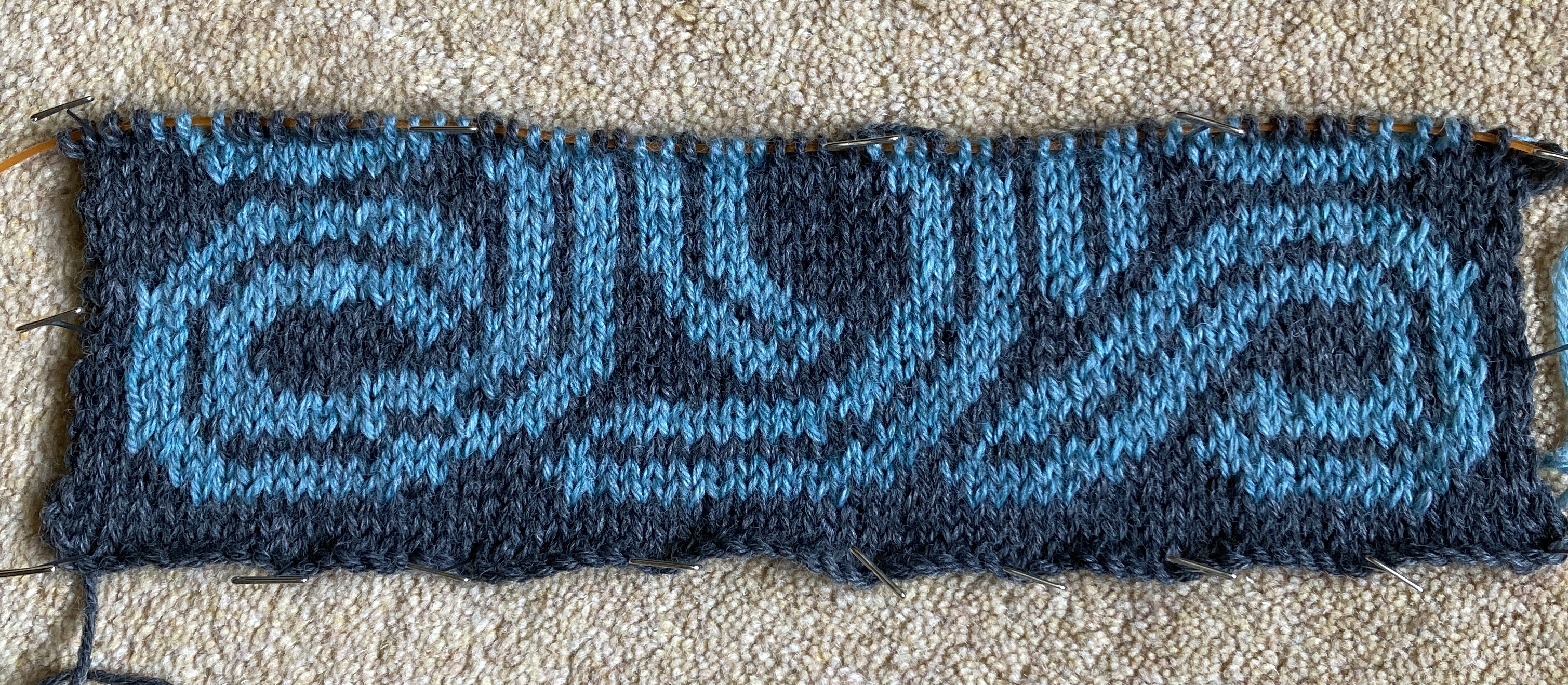 A piece of stranded knitting in progress. The background colour is dark grey and the pattern colour is blue. It shows the beginnings of a Celtic knot and is pinned onto a pale gold carpet.