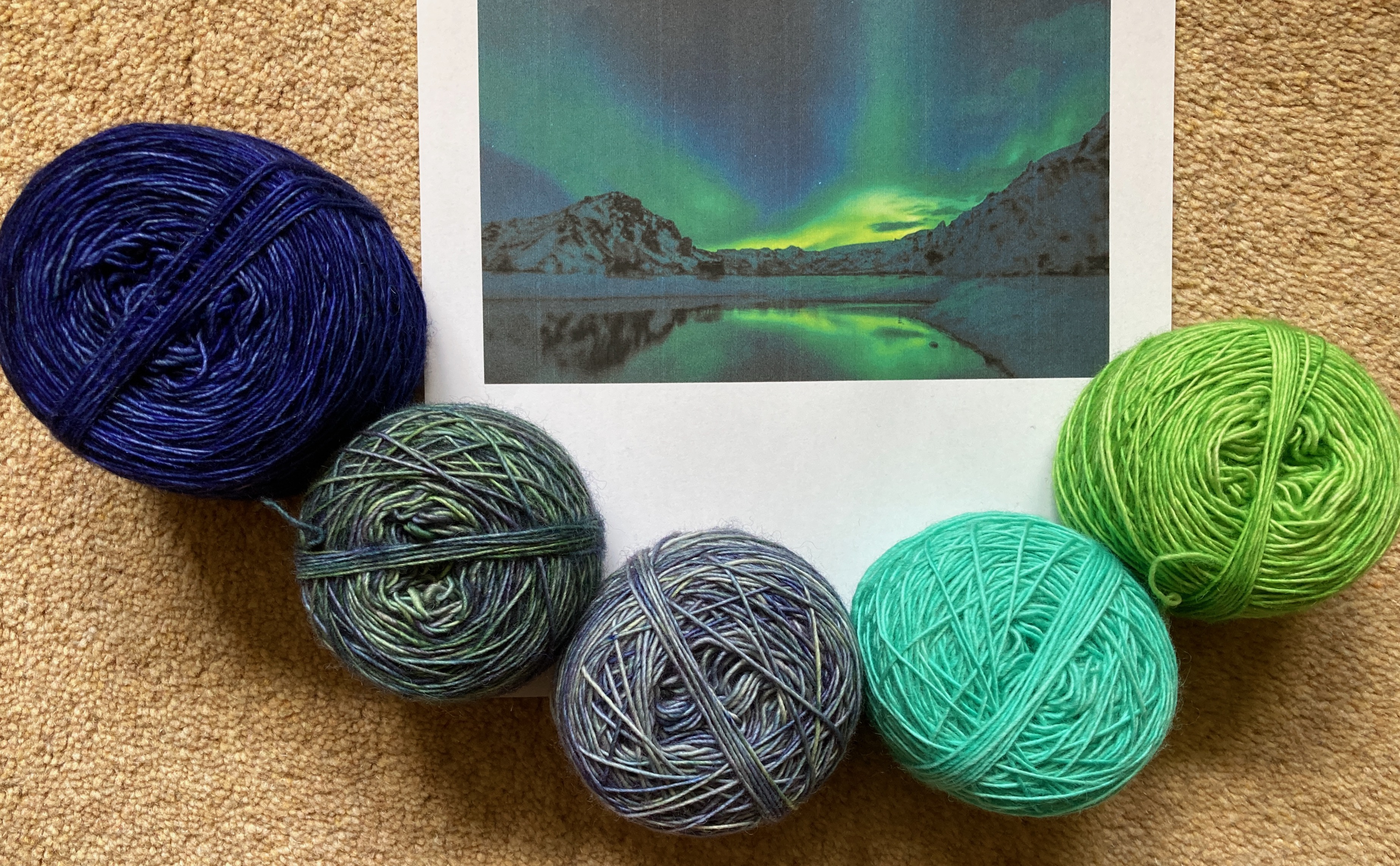 Five balls of yarn curve around a photo of the Northern Lights. The yarn matches the colours in the picture - dark blue, blue-green, grey, acid aqua and apple green. The background is a pale gold carpet