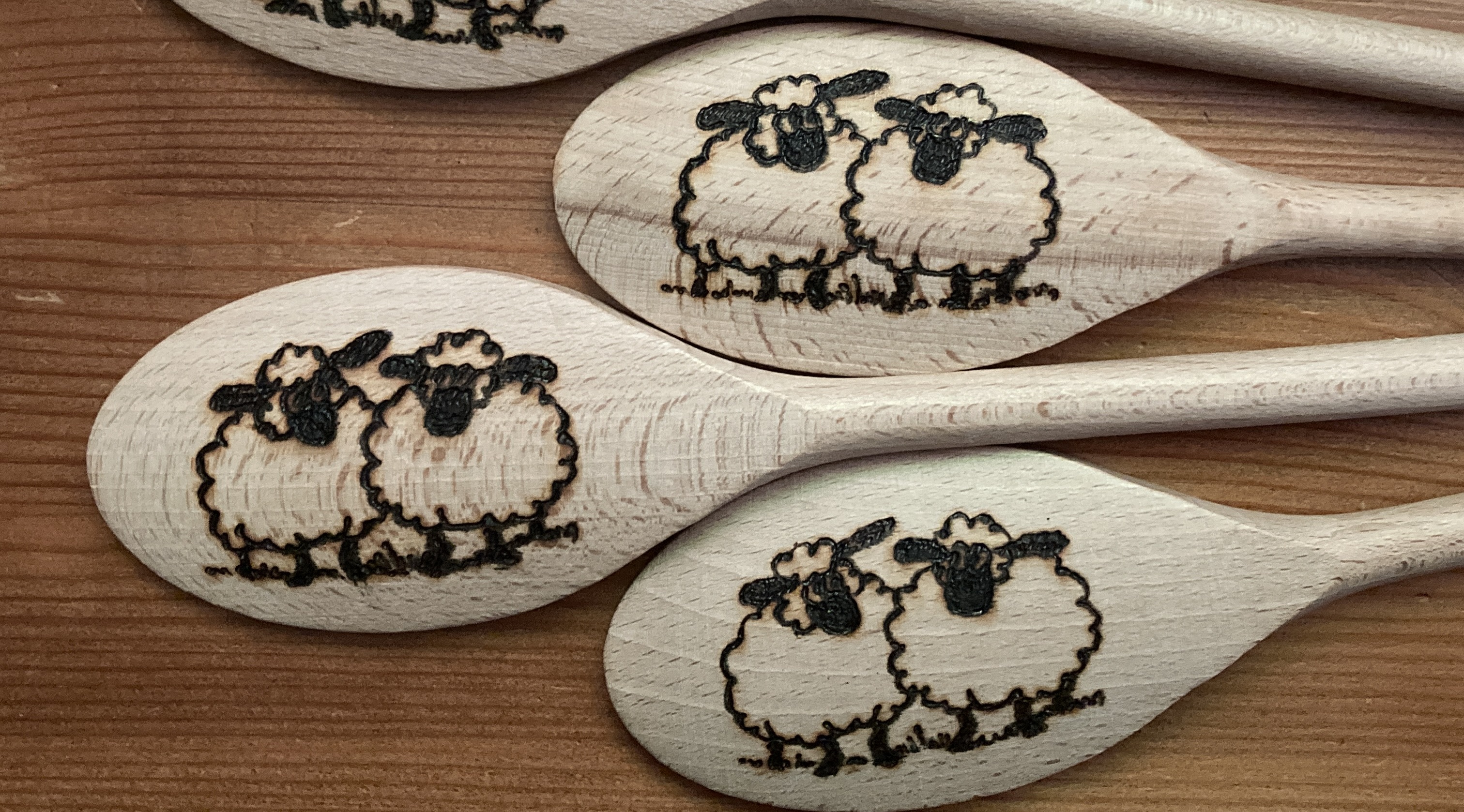 Three wooden spoons lie face down on a darker wooden desk. The back of each is decorated with a pyrography design of two comical sheep. A fourth spoon is just visible at the top of the image.