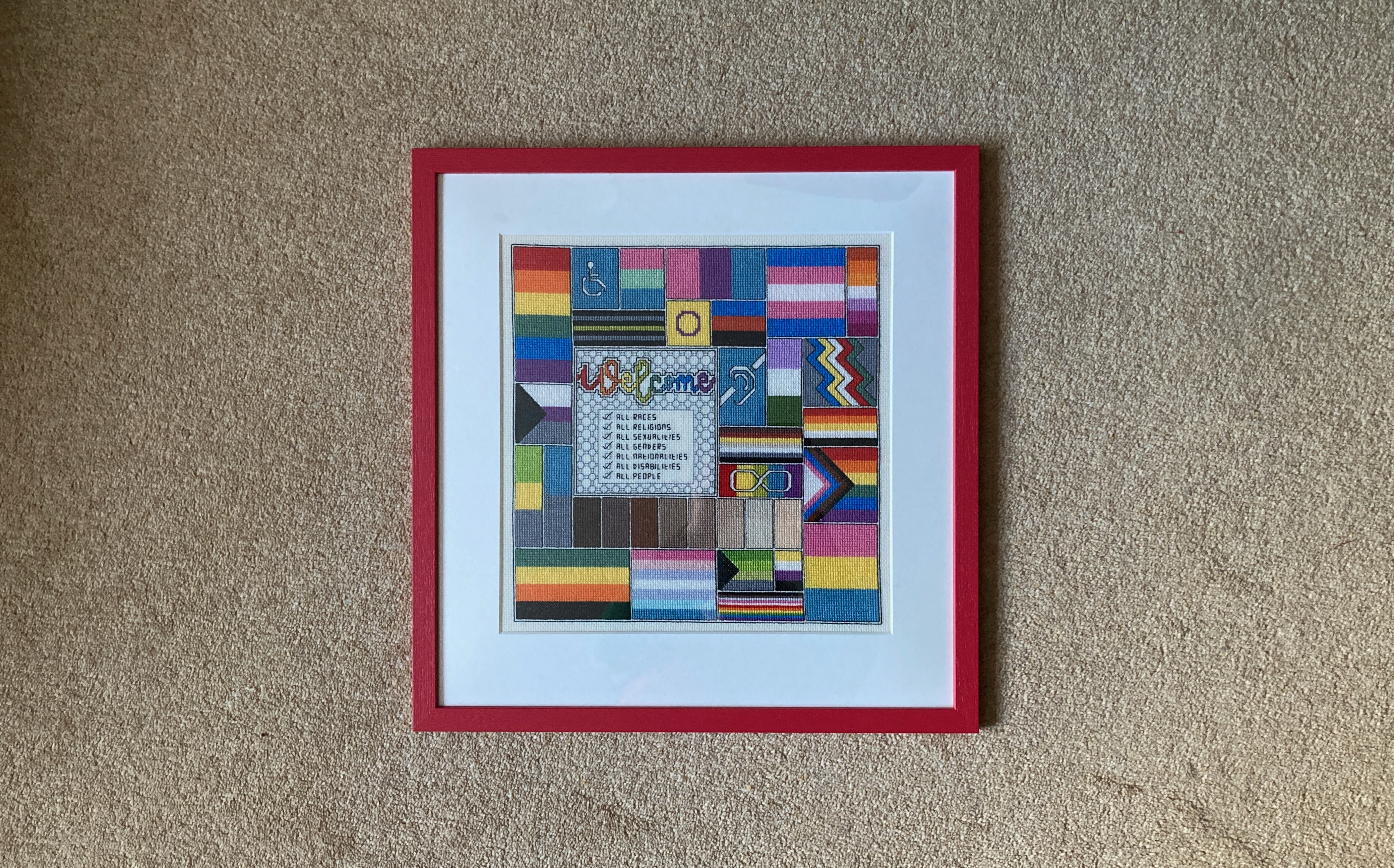 A cross-stitch montage of a wide variety of pride flags and skin tones for a brightly coloured square. Within that the words "Welcome All Races, All Religions, All Sexualities, All Genders, All Nationalities, All Disabilities, All People". A white mount surrounds the stitching with a narrow red frame. The background is a pale gold carpet.