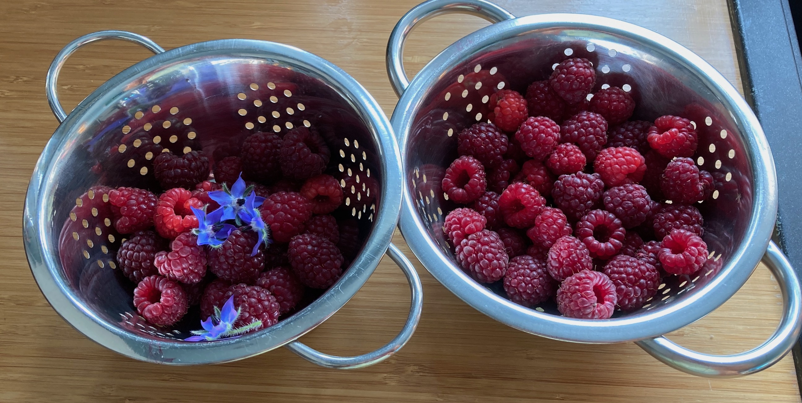 Two small metal colanders sit on a bamboo worktop, filled with raspberries. There are a few borage flowers in the one on the left as well.