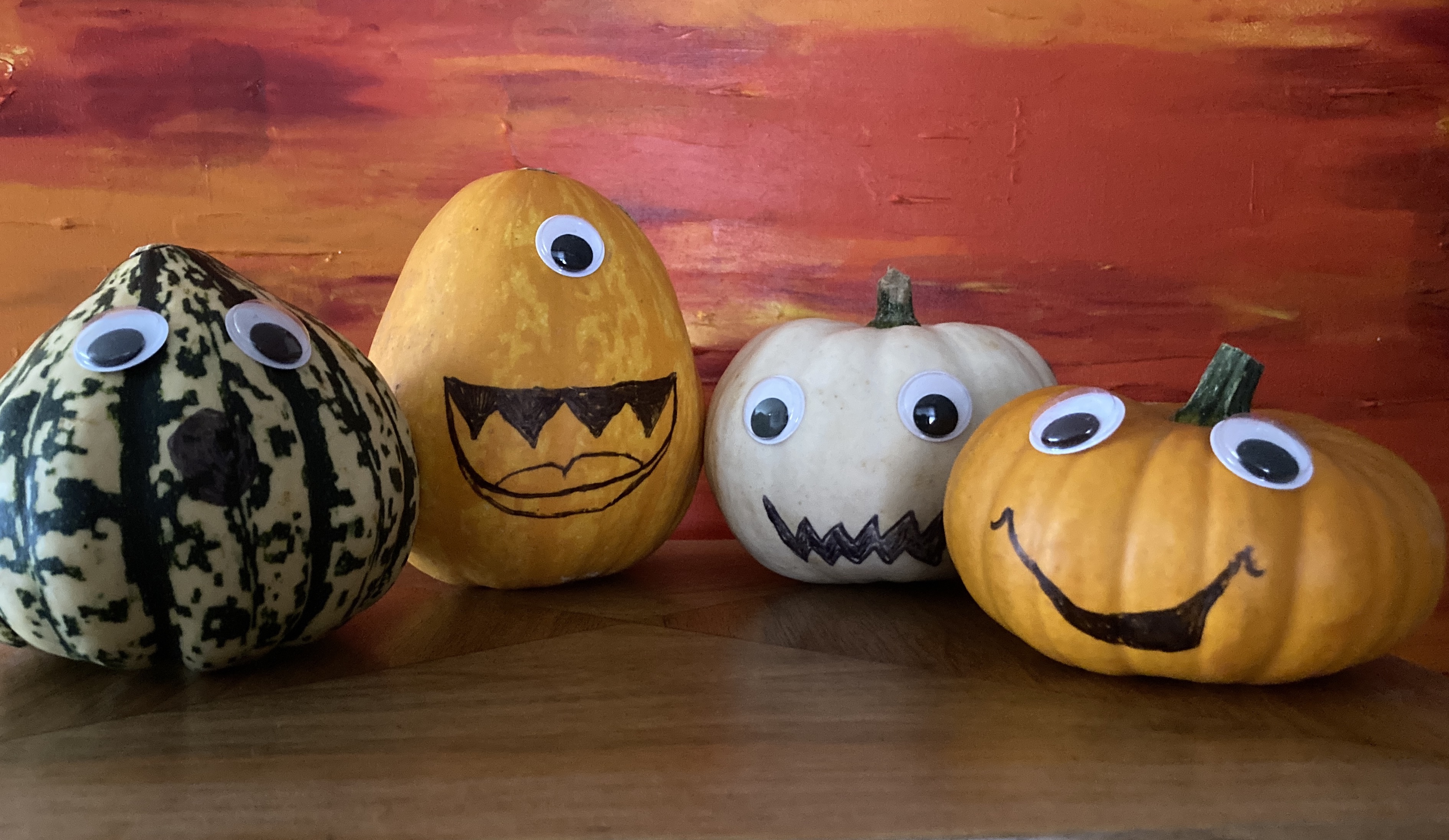 Four small pumpkins of different colours and shapes have googly eyes stuck on and mouths drawn with black pen sit on a wooden cabinet. Behind them is an acrylic painting in shades of orange and red.