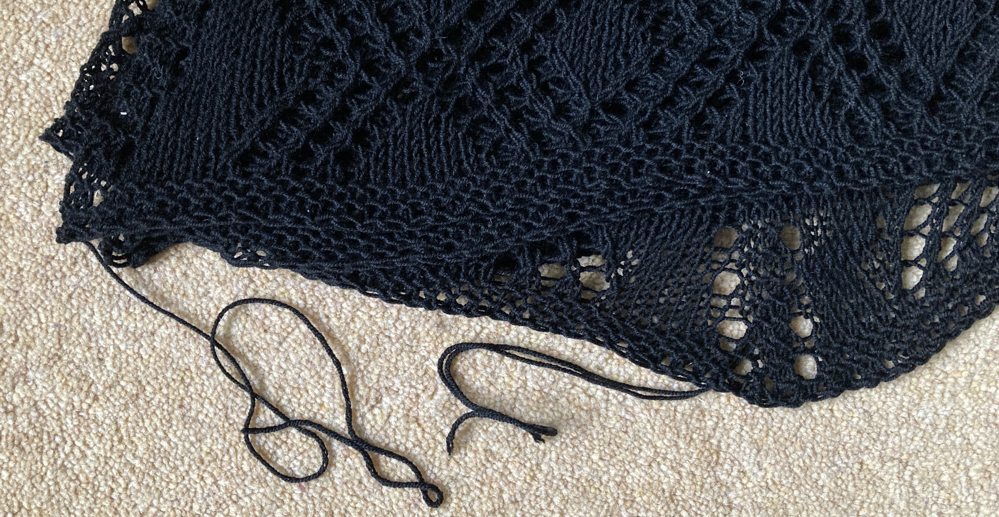 Part of a folded black lace knitted shawl on a pale carpet with yarn ends that need to be woven in