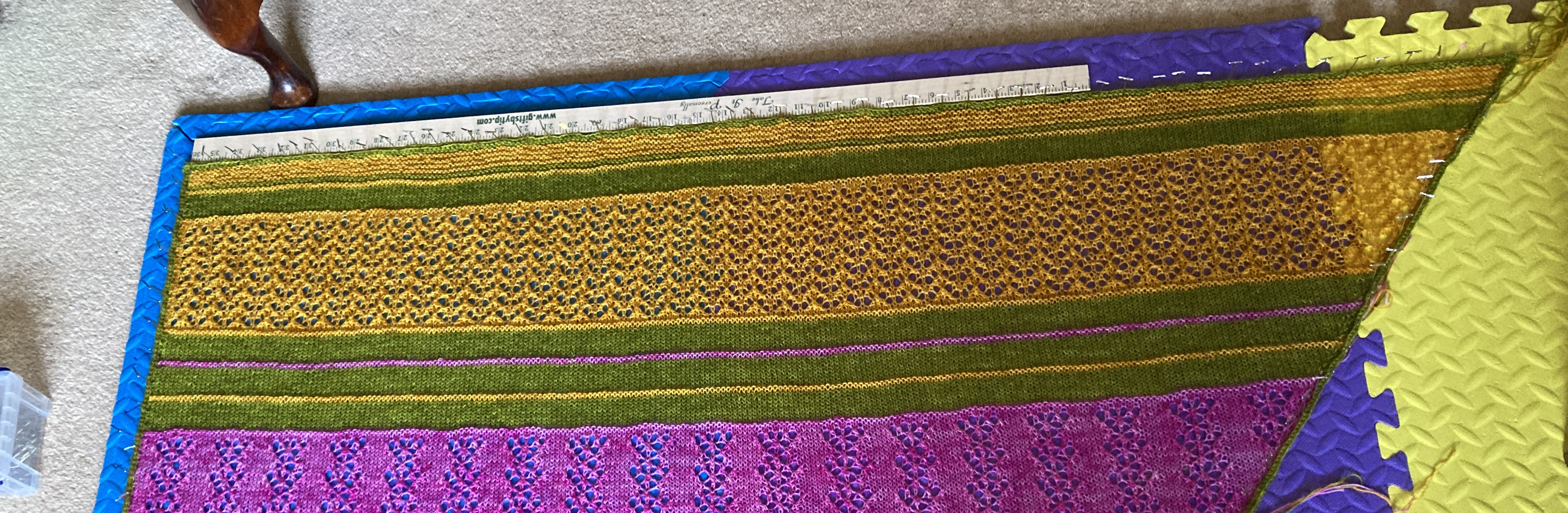 The top edge of a knitted shawl in golden yellow, green and pink, pinned out to dry with a wooden rule to keep the edge straight.