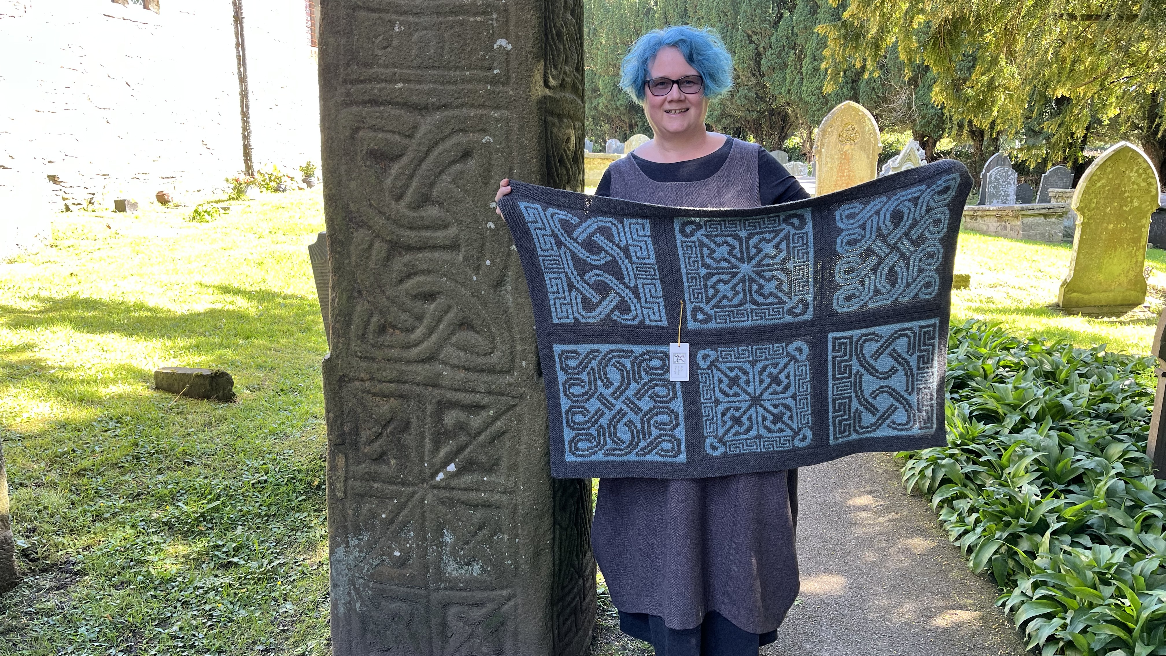 Me standing next to the Nevern Cross holding my Nevern Lap Blanket which uses stranded knitting designs created from the carved decoration on the cross.